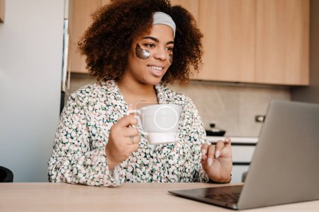 Téléchargez les photos : Jeune belle femme africaine souriante avec des patchs et une tasse de thé travaillant avec un ordinateur portable à la maison - en image libre de droit
