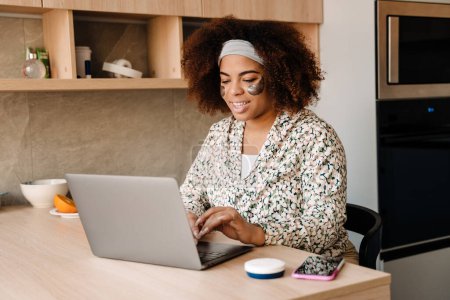 Téléchargez les photos : Jeune belle femme africaine souriante avec des patchs pour les yeux travaillant avec un ordinateur portable à la maison - en image libre de droit