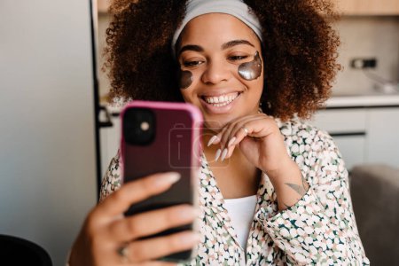 Téléchargez les photos : Jeune belle femme africaine souriante avec des patchs pour les yeux prenant selfie à la maison - en image libre de droit