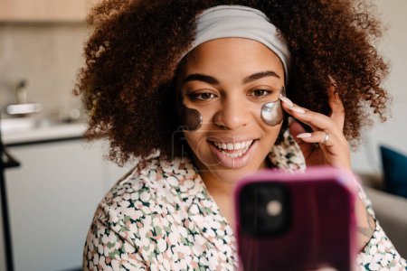 Photo for Young beautiful smiling african woman adjusting her eye patches while taking selfie at home - Royalty Free Image