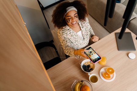 Téléchargez les photos : Vue du dessus de la belle femme afro-américaine souriante prenant en photo son petit déjeuner, flocons d'avoine aux fruits, jus d'orange et thé - en image libre de droit