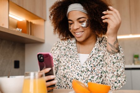 Téléchargez les photos : Jeune belle femme africaine souriante avec des patchs sous les yeux assis sur la cuisine avec un verre de jus d'orange et regardant son téléphone - en image libre de droit