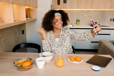 Photo for Young beautiful smiling african woman with under eye patches taking selfie during breakfast on cozy kitchen at home - Royalty Free Image