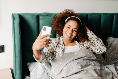Téléchargez les photos : Jeune femme afro-américaine souriante et utilisant un téléphone portable alors qu'elle était couchée dans son lit à la maison - en image libre de droit