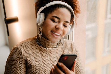 Téléchargez les photos : Jeune femme afro-américaine heureuse portant un casque souriant et utilisant un téléphone portable à la maison - en image libre de droit