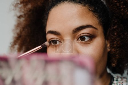 Téléchargez les photos : Jeune femme afro-américaine heureuse faisant maquillage par miroir à la maison - en image libre de droit