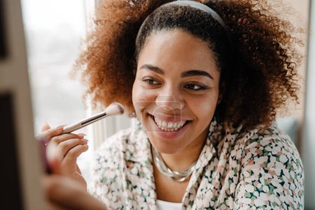 Téléchargez les photos : Jeune femme afro-américaine heureuse faisant maquillage par miroir à la maison - en image libre de droit