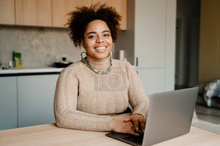 Téléchargez les photos : Jeune femme afro-américaine heureuse souriante et utilisant un ordinateur portable tout en étant assise sur la table à la maison - en image libre de droit
