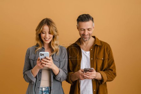 Téléchargez les photos : Blanc couple heureux à l'aide de téléphones mobiles tout en riant isolé sur fond beige - en image libre de droit