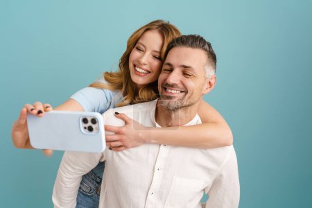 Téléchargez les photos : Blanc couple heureux prenant selfie photo sur téléphone portable tout en riant isolé sur fond bleu - en image libre de droit