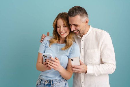 Foto de Blanco feliz pareja usando teléfonos móviles mientras se ríe aislado sobre fondo azul - Imagen libre de derechos