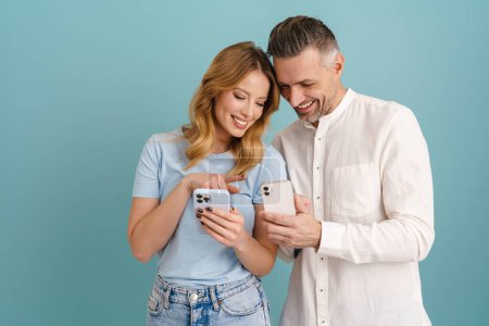 Téléchargez les photos : Blanc couple heureux à l'aide de téléphones mobiles tout en riant isolé sur fond bleu - en image libre de droit