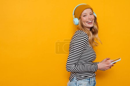Foto de Mujer rubia joven usando el teléfono celular mientras escucha música aislada sobre fondo amarillo - Imagen libre de derechos
