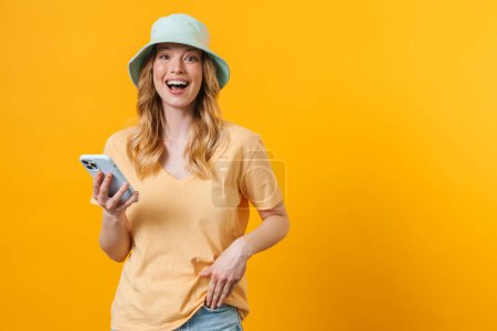 Téléchargez les photos : Jeune femme blonde portant panama riant et utilisant un téléphone portable isolé sur fond jaune - en image libre de droit