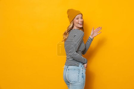 Foto de Mujer rubia joven con sombrero sonriendo y haciendo gestos a la cámara aislada sobre fondo amarillo - Imagen libre de derechos