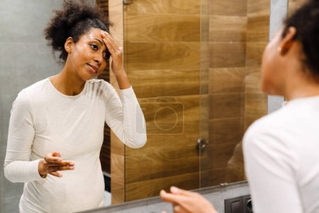 Photo for Young beautiful pregnant afro woman looks in the mirror and smearing her face with cream at home - Royalty Free Image
