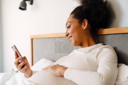 Foto de Young smiling beautiful pregnant afro woman liying on bed and looking on her phone in cozy bedroom at home - Imagen libre de derechos