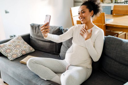 Photo for Young beautiful smiling happy pregnant afro woman sitting on sofa touching her chest and taking selfie on phone in cozy room at home - Royalty Free Image