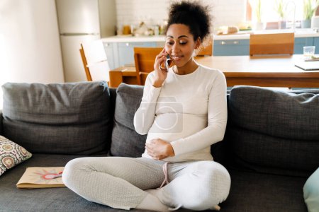 Young beautiful smiling happy pregnant afro woman sitting on sofa holding her belly and talking on phone in cozy room at home