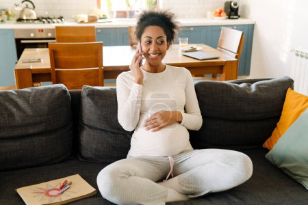 Photo for Young beautiful smiling happy pregnant afro woman sitting on sofa and talking on her phone in cozy room at home - Royalty Free Image