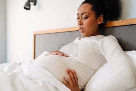Téléchargez les photos : Adult beautiful pregnant tense african woman holding her belly while liying on bed - en image libre de droit