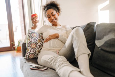 Adult beautiful pregnant smiling african woman eating apple and holding her belly while sitting on sofa at home