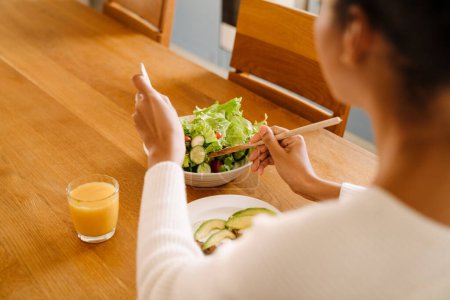 Téléchargez les photos : Back view of african woman eating salad with glass of orange juice close-up in cozy kitchen at home - en image libre de droit