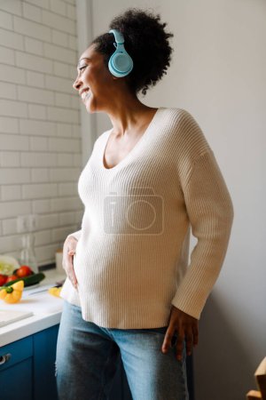 Photo for Adult beautiful happy smiling pregnant african woman in headphones holding her belly and standing in cozy kitchen - Royalty Free Image