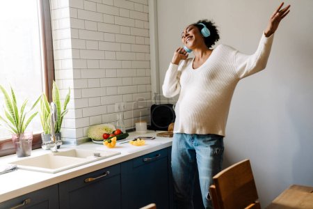Téléchargez les photos : Adult beautiful happy smiling pregnant african woman in headphones dancing with closed eyes while cooking in cozy kitchen - en image libre de droit