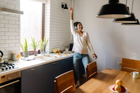Téléchargez les photos : Adult beautiful happy smiling pregnant african woman in headphones dancing with closed eyes while cooking in cozy kitchen - en image libre de droit