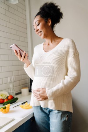 Téléchargez les photos : Adult beautiful smiling african pregnant woman looking on her phone and holding her belly while cooking salad in cozy kitchen at home - en image libre de droit