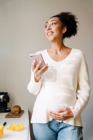 Téléchargez les photos : Adult beautiful smiling african pregnant woman with phone holding her belly while standing in cozy kitchen at home - en image libre de droit