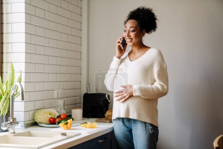 Foto de Adult beautiful smiling african pregnant woman talking phone and holding her belly while cooking salad in cozy kitchen at home - Imagen libre de derechos