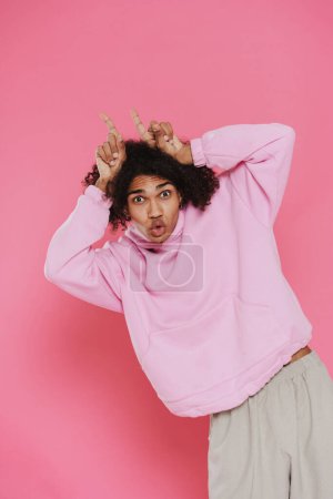 Téléchargez les photos : Young handsome stylish latin curly grimacing man showing horns and looking at camera while standing over isolated pink background - en image libre de droit