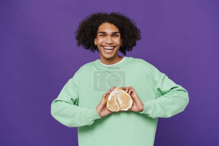 Foto de Young smiling happy latin man holding pomelo on his chest and looking at camera while standing over isolated violet background - Imagen libre de derechos