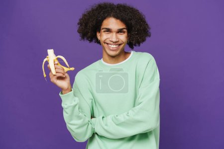 Photo for Young smiling handsome latin man holding bitened banana in hand and looking at camera while standing over isolated violet background - Royalty Free Image