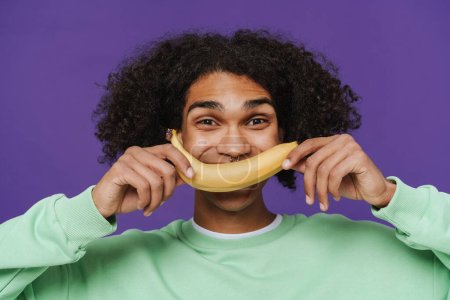 Téléchargez les photos : Young curly latin man holding banana on face like smile while standing over isolated violet background - en image libre de droit