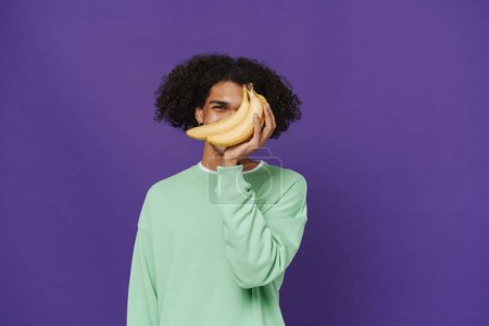 Photo for Young latin man covering his face with bunch of bananas and looking at camera with one eye, while standing over isolated violet background - Royalty Free Image