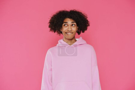 Photo for Young caribbean man with piercing looking aside while biting his lip isolated over pink background - Royalty Free Image