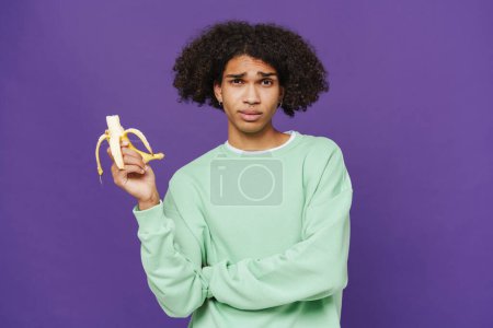 Photo for Young caribbean man frowning while eating banana isolated over purple background - Royalty Free Image