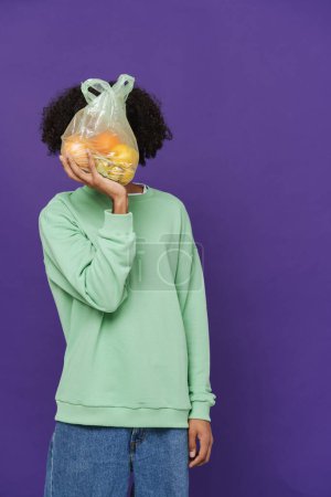 Photo for Young caribbean man holding and showing plastic bag with fruits isolated over purple background - Royalty Free Image