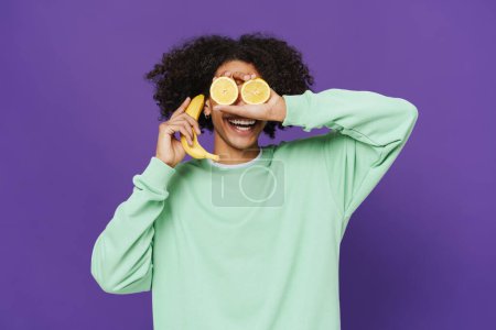 Photo for Young caribbean man smiling while making fun with fruits isolated over purple background - Royalty Free Image