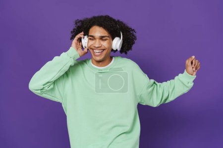Téléchargez les photos : Young caribbean man dancing while listening music with headphones isolated over purple background - en image libre de droit