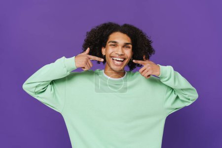 Téléchargez les photos : Young caribbean man with piercing pointing fingers at his smile isolated over purple background - en image libre de droit