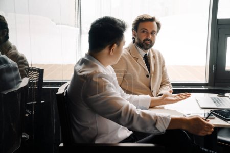 Téléchargez les photos : Adult multiracial businessmen talking and using laptop while sitting by table indoors - en image libre de droit