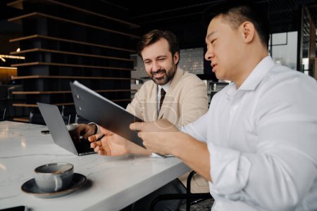 Foto de Asian businessman showing documents to his smiling partner with laptop and discussing working moments sitting at a table in a modern office - Imagen libre de derechos