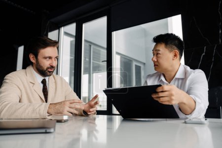 Téléchargez les photos : Adult businessman discussing with asian partner who holding clip board with plan, while they sitting at modern office building - en image libre de droit