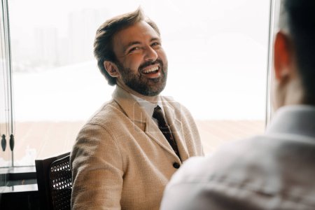 Téléchargez les photos : Adult white businessman smiling while sitting with his male partner by table indoors - en image libre de droit