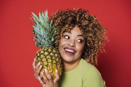 Téléchargez les photos : Young black woman laughing while posing with pineapple isolated over red background - en image libre de droit