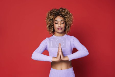 Foto de Young black woman with afro curls holding palms together while meditating isolated over red background - Imagen libre de derechos